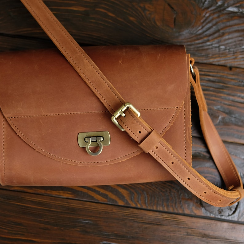 May include: A brown leather satchel bag with a flap closure and a brass buckle. The bag has a long adjustable strap.