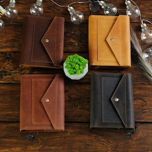 May include: Four leather journals in various colors: dark brown, tan, medium brown, and black. Each journal features a unique envelope-style flap closure with a silver snap. The journals are arranged on a dark wooden surface.