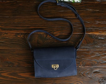 Puede incluir: Un bolso bandolera de cuero azul oscuro con cierre dorado y una correa larga. El bolso está sobre una superficie de madera marrón oscura.