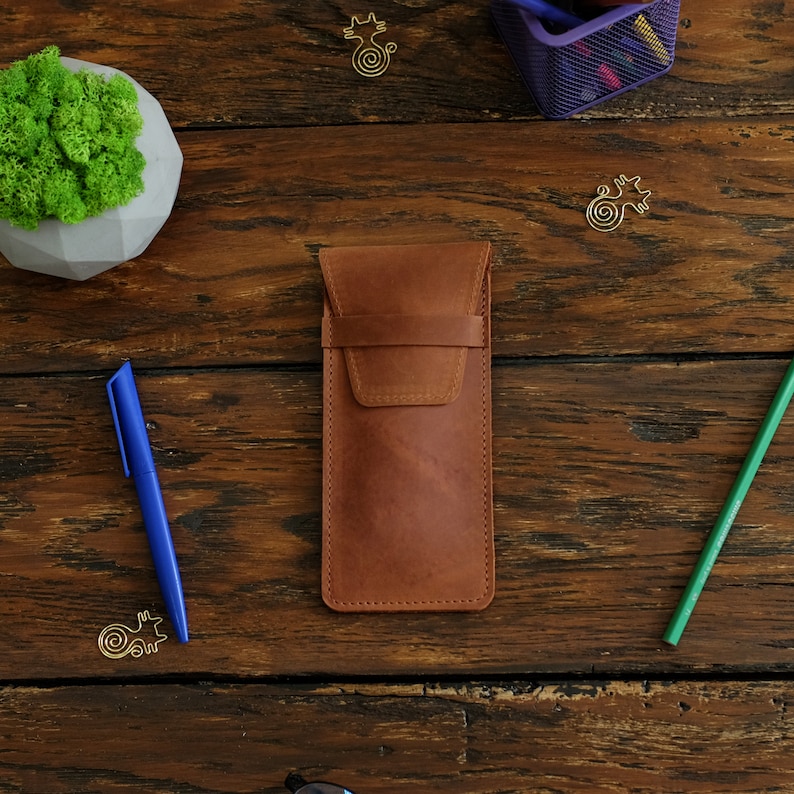 Op de afbeelding: Een bruine leren potlood etui met een klep sluiting. De etui ligt op een houten oppervlak met een blauwe pen en een groen potlood.