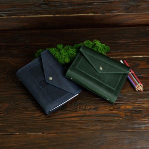 May include: Two leather document holders, one navy blue and one forest green, with a flap closure and a silver snap. The green holder has the name "Oliver" embossed on it. A bundle of colored pencils rests nearby.