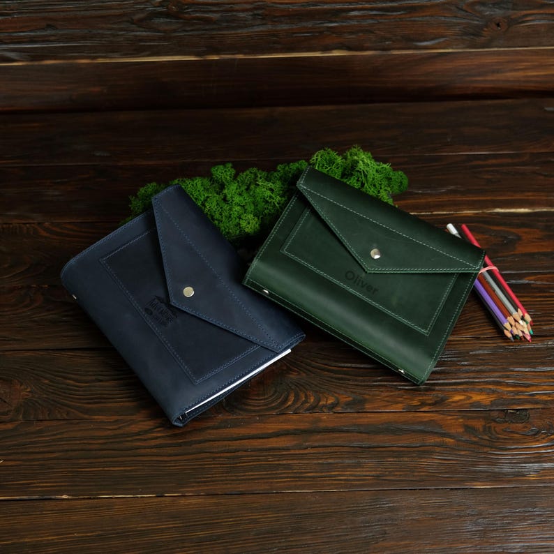 May include: Two leather document holders, one navy blue and one forest green, with a flap closure and a silver snap. The green holder has the name "Oliver" embossed on it. A bundle of colored pencils rests nearby.