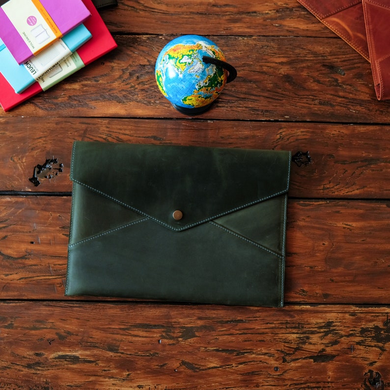 May include: A dark green leather envelope style clutch with a brass button closure. The clutch is on a wooden surface with a globe and notebooks.
