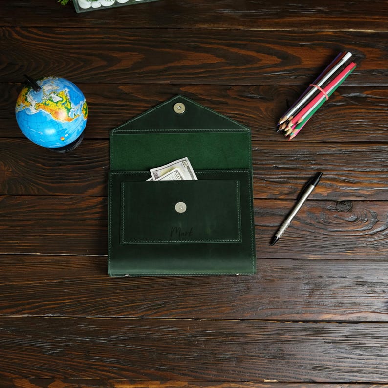 May include: A dark green leather envelope-style wallet with a flap, open to reveal a pocket holding cash. A small silver button secures the flap. A globe, colored pencils, and a pen are also on the wooden surface.