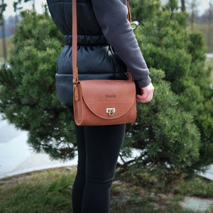 May include: A brown leather crossbody bag with a gold clasp and the name "Emily" engraved on the front.