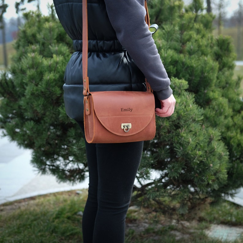 May include: A brown leather crossbody bag with a gold clasp and the name "Emily" engraved on the front.