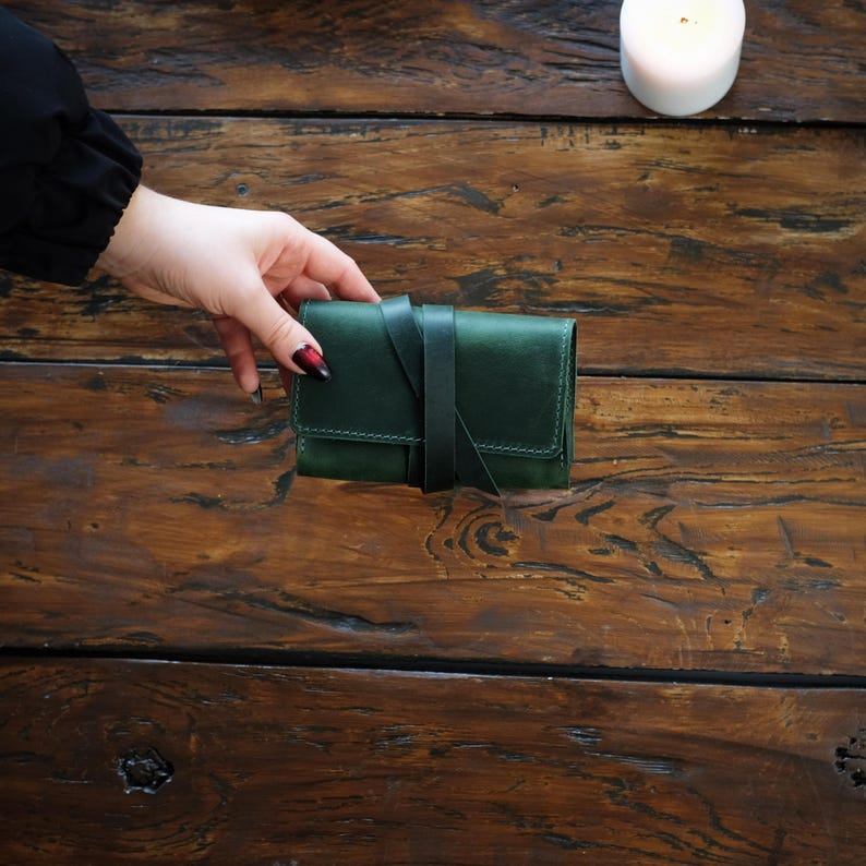 Pode incluir: Uma carteira de couro verde-escura de formato retangular com costuras vis&iacute;veis ao longo das bordas e uma tira de couro a condizer que envolve o centro para prender o fecho de aba, com um acabamento liso e mate em toda a pe&ccedil;a.