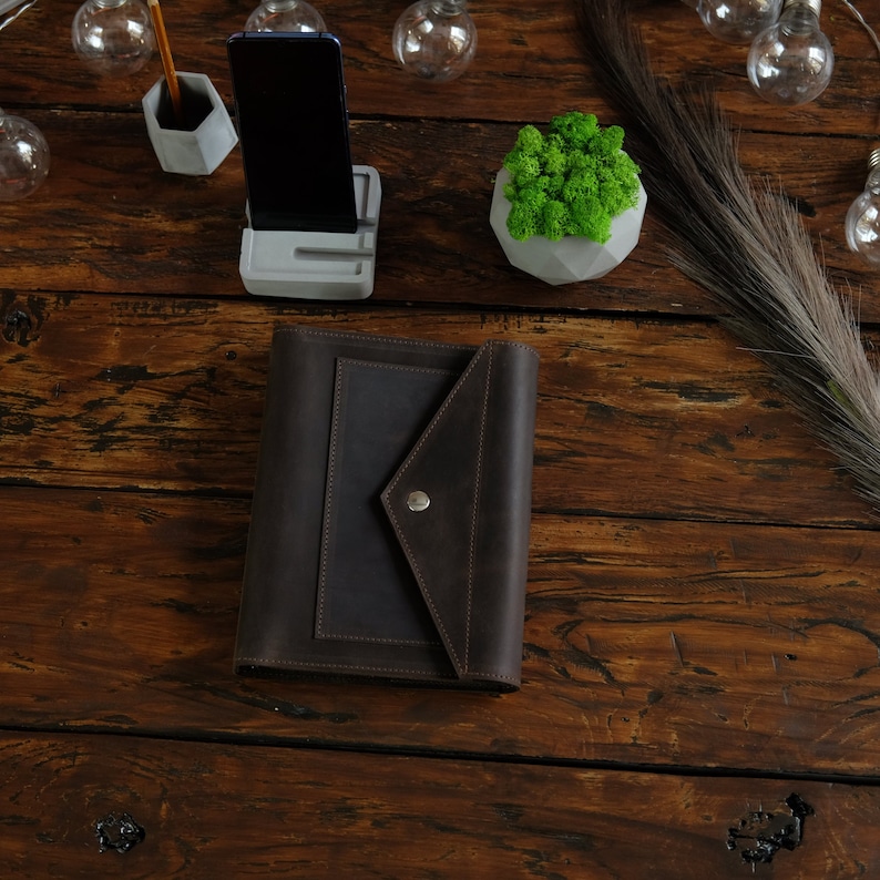 May include: A brown leather journal with a flap closure and a metal button. The journal is on a wooden table with a small potted plant, a phone stand, and a pencil holder.