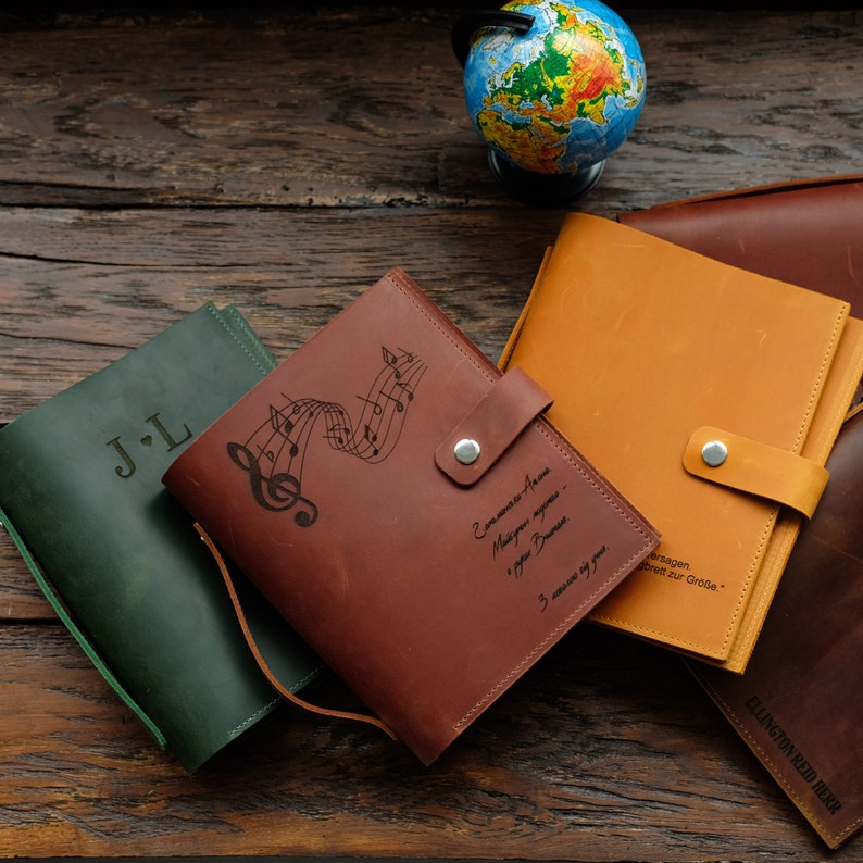 May include: Four leather-bound journals with different personalized engravings. The journals are brown, green, orange, and a darker brown. The journals have a flap closure with a button. The brown journal has a musical note design and the text "Eine Melodie f&uuml;r Anna - Musik f&uuml;r meine eigene Welt -  J.L.  3 Melodien f&uuml;r dich" engraved on the front.