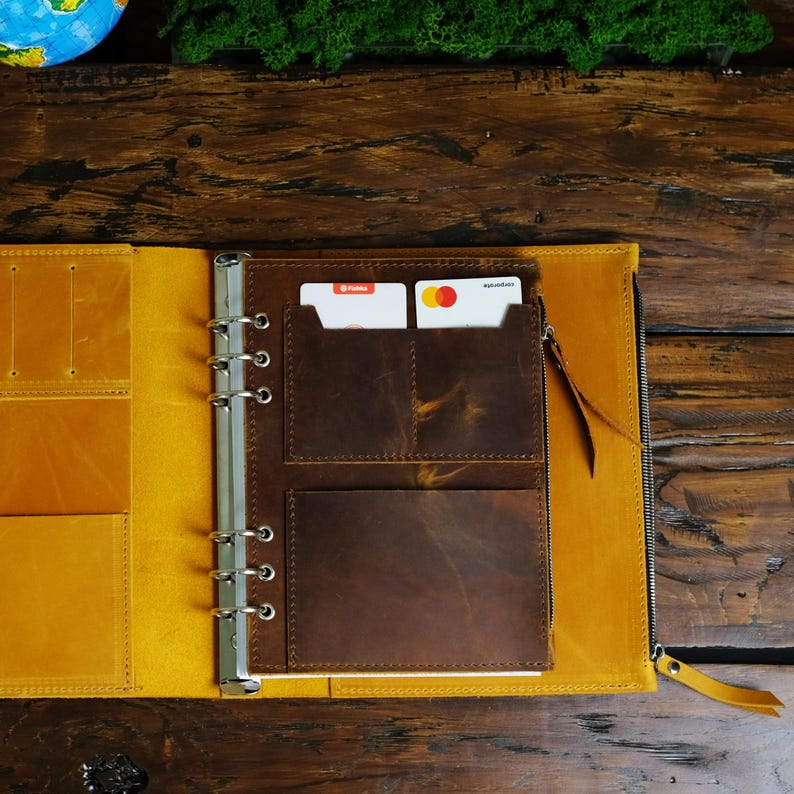 May include: Open leather binder with a yellow exterior and brown interior pockets. The binder has a zipper closure and a silver ring mechanism. Two credit cards are visible in a pocket.
