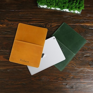 Könnte beinhalten: Zwei Lederhüllen für Tablets, eine gelbe und eine grüne, mit dem Namen "Robert" auf der gelben Hülle geprägt. Ein weißes Tablet liegt auf den Hüllen.