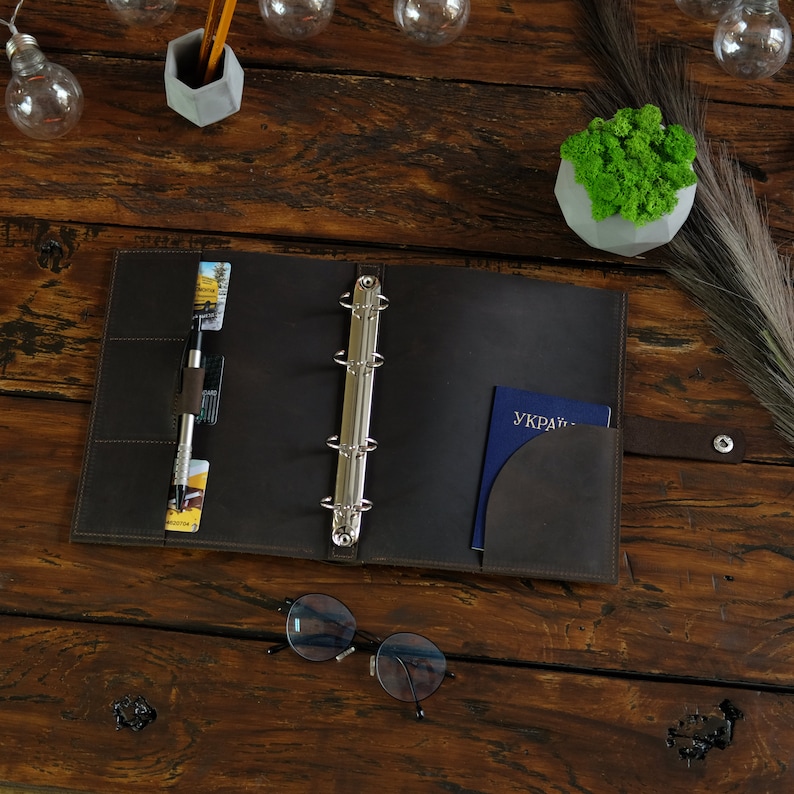 May include: A brown leather binder with a silver ring mechanism. The binder has a pocket on the right side and a pen and cards are inserted in the left pocket. The binder is open and a blue passport with the word "UKRAINE" is visible in the right pocket.