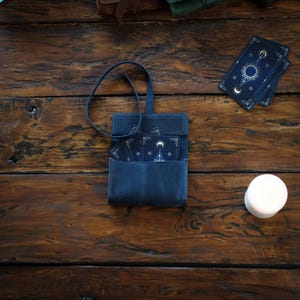 Pode incluir: Uma bolsa de couro azul-escura com uma al&ccedil;a longa contendo um baralho com desenhos celestiais de sol, lua e estrelas. As cartas s&atilde;o pretas com linhas douradas. Uma vela branca est&aacute; posicionada &agrave; direita da bolsa.