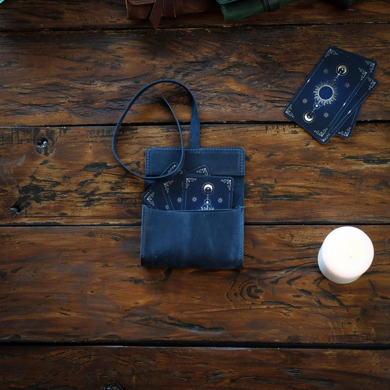 Pode incluir: Uma bolsa de couro azul-escura com uma al&ccedil;a longa contendo um baralho com desenhos celestiais de sol, lua e estrelas. As cartas s&atilde;o pretas com linhas douradas. Uma vela branca est&aacute; posicionada &agrave; direita da bolsa.