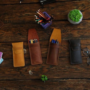 Op de afbeelding: Vier leren potloodkokers, twee bruin, &eacute;&eacute;n beige en &eacute;&eacute;n zwart, met verschillende schrijfbenodigdheden erin. De kokers liggen op een houten oppervlak.