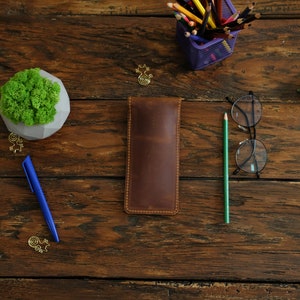 Op de afbeelding: Een bruine leren potlood etui op een houten tafel met een blauwe pen, een kleine plant in een pot, een bril en een houder met kleurpotloden.