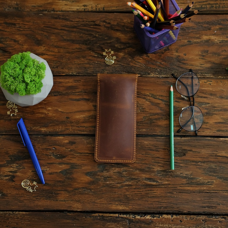 Op de afbeelding: Een bruine leren potlood etui op een houten tafel met een blauwe pen, een kleine plant in een pot, een bril en een houder met kleurpotloden.