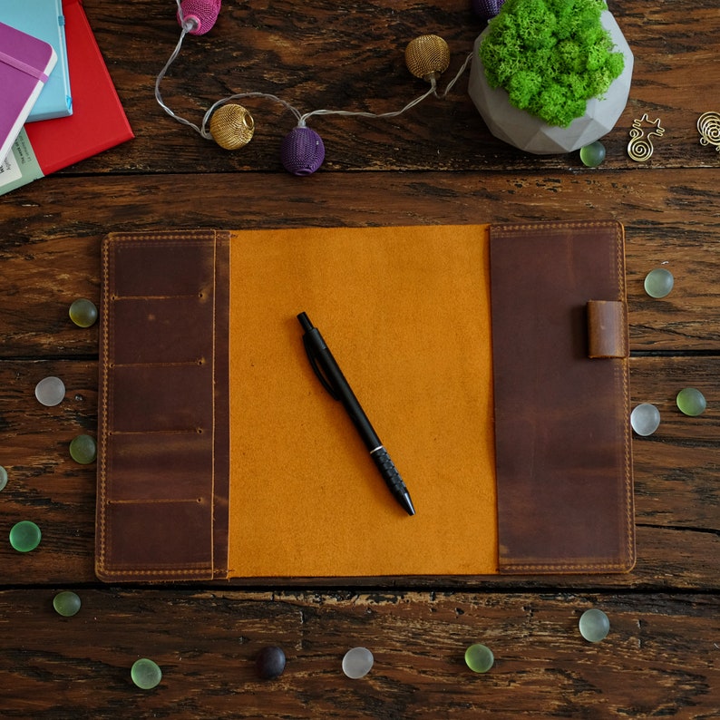 Puede incluir: Un diario de cuero marr&oacute;n con un inserto de fieltro amarillo y un bol&iacute;grafo negro. El diario est&aacute; abierto y acostado sobre una superficie de madera. Hay peque&ntilde;os objetos decorativos esparcidos alrededor del diario.