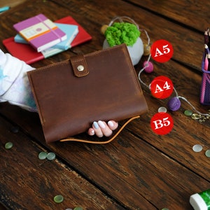 Könnte beinhalten: Ein braunes Ledernotizbuch mit einem Metallverschluss und einem braunen Lederriemen. Das Notizbuch wird von einer Hand mit weißem Nagellack gehalten. Das Notizbuch liegt auf einem Holztisch mit einem Stapel Notizbücher und einer kleinen grünen Pflanze in einem Topf.