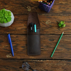 Op de afbeelding: Een bruine leren potloodhouder met drie zwarte pennen en een turquoise markeerstift erin. De houder ligt op een houten oppervlak.