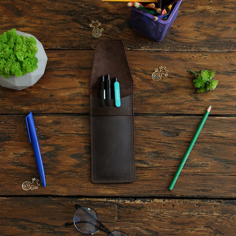 Op de afbeelding: Een bruine leren potloodhouder met drie zwarte pennen en een turquoise markeerstift erin. De houder ligt op een houten oppervlak.