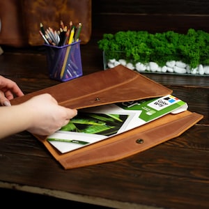 Puede incluir: Una carpeta de documentos de cuero marrón estilo sobre con cierre a presión, que contiene un paquete de papel Color Way con la imagen de una serpiente verde. La carpeta está hecha de cuero liso con acabado mate y costuras visibles en los bordes.
