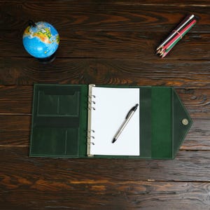 May include: An open, dark green leather binder with a blank white notepad and a silver pen. A small blue globe and a bundle of colored pencils are also on the dark wooden surface.
