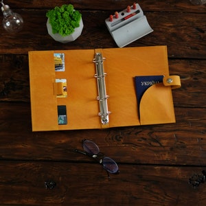 May include: A yellow leather binder with a silver ring mechanism. The binder has pockets for cards and a flap for a passport. The passport has the word "UKRAINE" printed on it.