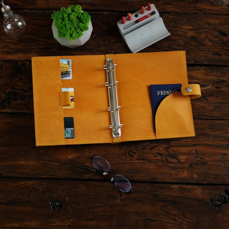 May include: A yellow leather binder with a silver ring mechanism. The binder has pockets for cards and a flap for a passport. The passport has the word "UKRAINE" printed on it.