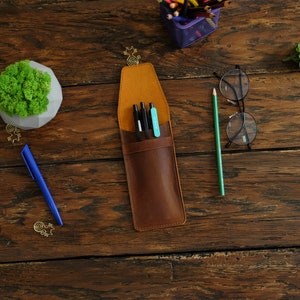 Op de afbeelding: Een bruin leren potlood etui met een klep sluiting. De etui is open en heeft drie zwarte pennen en een blauwe pen erin. De etui ligt op een houten oppervlak.