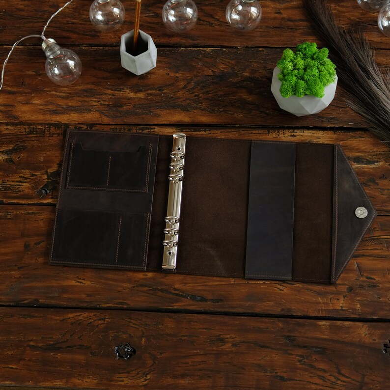May include: A brown leather bound planner with a silver ring binder and a flap closure. The planner is open and lying on a wooden surface.