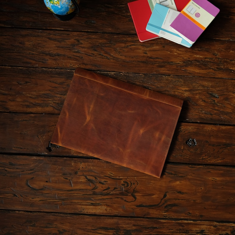 May include: A brown leather laptop sleeve on a wooden surface.