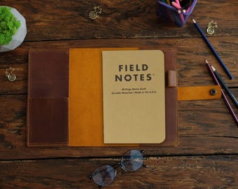 May include: A brown leather notebook cover with a tan interior, featuring a Field Notes 48-page memo book. The cover includes a side pocket, a pen loop, and a snap button closure strap. The notebook cover is open, displaying the tan memo book.