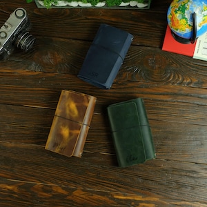 May include: Three leather journals in navy, brown, and green, each secured with an elastic band. The navy journal is embossed with "JSR", the brown journal with "Mark", and the green journal with "Oliver". The journals are on a wooden surface.