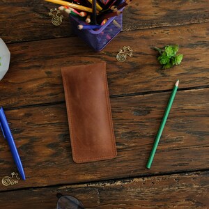Op de afbeelding: Een bruine leren potlood etui op een houten oppervlak. Het etui is gesloten en heeft een gestikte rand. Een groen potlood en een blauwe pen zijn ook zichtbaar.