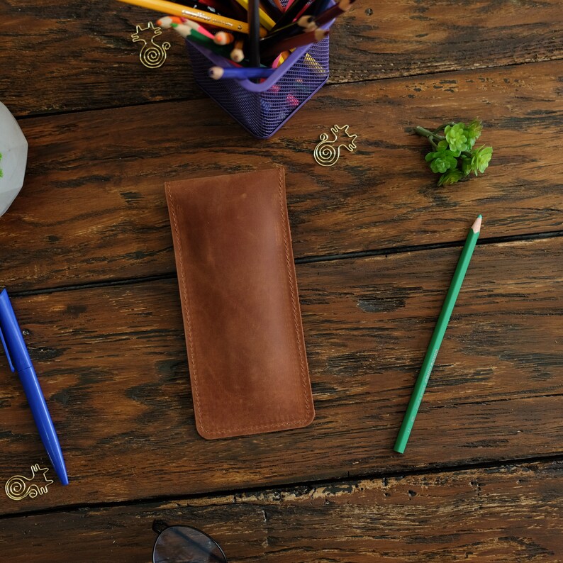 Op de afbeelding: Een bruine leren potlood etui op een houten oppervlak. Het etui is gesloten en heeft een gestikte rand. Een groen potlood en een blauwe pen zijn ook zichtbaar.