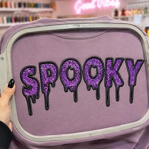 May include: A mauve sweatshirt secured in a rectangular embroidery hoop. The fabric features the word SPOOKY embroidered in a dripping font style with purple glitter material outlined in thick black thread, creating a textured appearance.