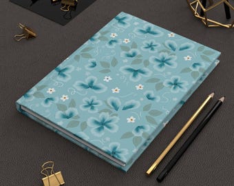 May include: A rectangular notebook with a light blue cover featuring a repeating pattern of dark blue flowers, small white blossoms, and green leaves. The notebook is accompanied by one gold-toned pen, one black pen, and several gold-toned metal binder clips.