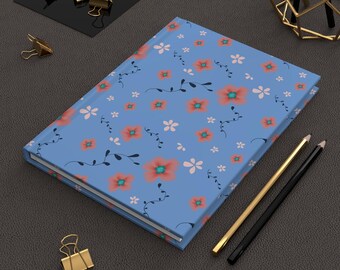 May include: A floral-patterned journal with a light blue cover. The design features orange and white flowers with green centers, and black vines. Gold and black pencils and gold binder clips are also visible.