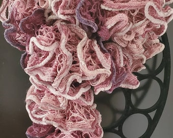 May include: A handmade, ruffled scarf in shades of pink, mauve, and white. The scarf has a delicate, open-weave design with a textured, layered appearance. The scarf is hanging on a black metal decorative ring.