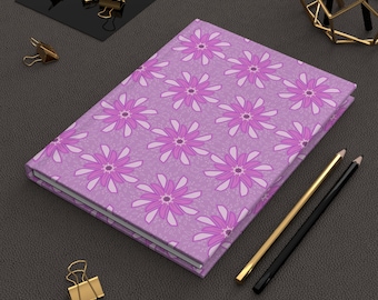 May include: A closed journal with a floral pattern in shades of purple and white. The journal is surrounded by gold and black office supplies, including binder clips and pencils. The cover design features repeating stylized flowers.