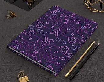 May include: A closed journal with a dark blue cover featuring a repeating pattern of abstract shapes in shades of purple and pink. Gold and black pencils and gold binder clips are arranged around the journal.