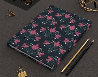 May include: A floral patterned journal with a dark blue cover. The design features pink flowers, green leaves, and white accents. Gold and black pencils and gold binder clips are also visible.