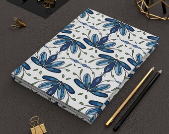 May include: A rectangular notebook featuring a white cover with a repeating pattern of blue dragonfly illustrations, thin green stems, and small dots. The notebook is accompanied by a gold-toned pen, a black pen, and several gold-toned metal binder clips.