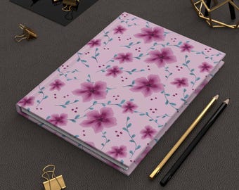 May include: A hardcover journal with a light pink cover featuring a floral pattern of purple flowers and green vines. Two pencils, one gold and one black, are next to the journal. Gold binder clips are scattered around.