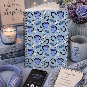 May include: A spiral-bound notebook with a light blue cover featuring a repeating pattern of purple mugs, black headphones, and white rectangular icons. The notebook is positioned upright, displaying a consistent graphic design across its front surface.