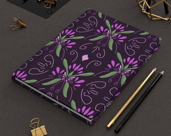 May include: A hardcover journal with a dark purple cover featuring a floral pattern in shades of purple, green, and white. The journal is open, revealing blank pages. Gold and black pencils and gold binder clips are nearby.
