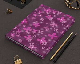 May include: A rectangular notebook with a deep purple cover featuring a repeating pattern of bright pink and light purple flowers with thin stems. The notebook is accompanied by a gold-colored pencil, a black pencil, and several gold-colored binder clips.