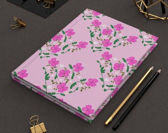 May include: A closed journal with a pink cover featuring a floral pattern of pink flowers, green leaves, and white blossoms. Gold and black pencils and gold binder clips are on the dark surface.