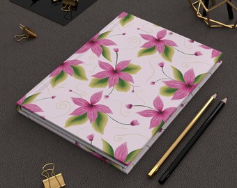 May include: A hardcover notebook with a light pink cover featuring a repeating pattern of pink five-petaled flowers with green leaves and thin gold swirling lines. The notebook is positioned next to a gold pencil and a black pencil.